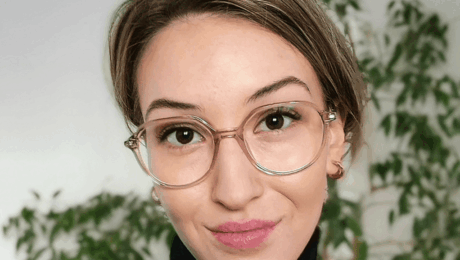 Kwadrat na strone (7) Woman with short hair and round glasses, wearing a black turtleneck, smiling softly indoors with green plants behind her